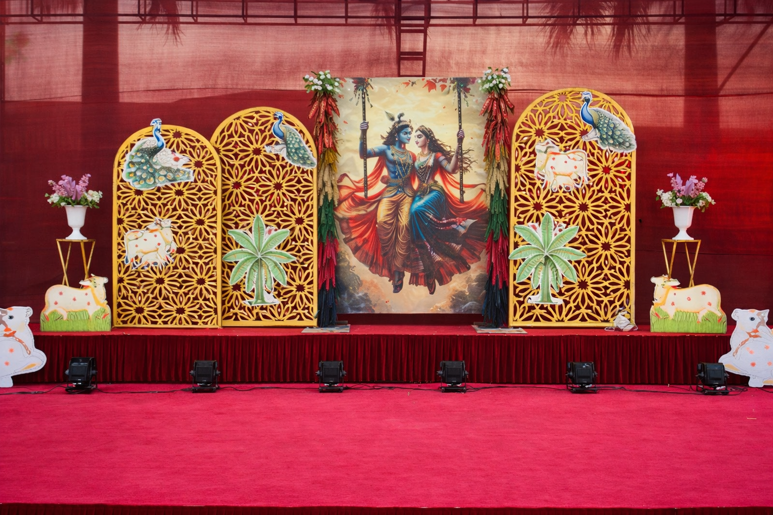 Radha Krishna theme wedding stage decoration with traditional village backdrop, golden floral panels, peacock cutouts, cow props, palm tree elements and elegant red carpet setup by Jatin Flower Mart. Perfect for Garba night, cultural events, wedding functions and festive celebrations.